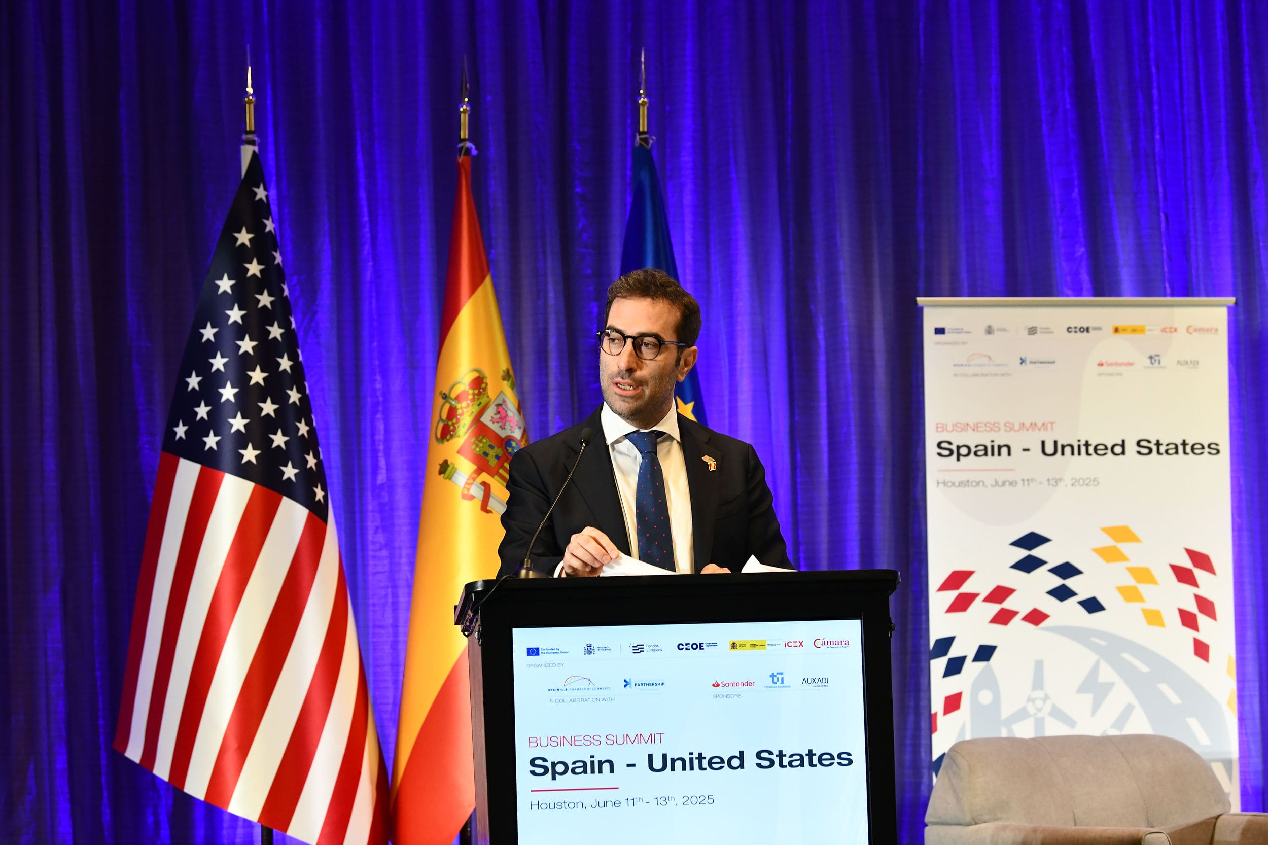 persona en un atril hablando y la bandera de Estados Unidos y de España