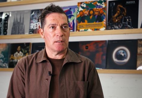 Hombre moreno conversa delante de una estantería con una colección de discos de vinilo
