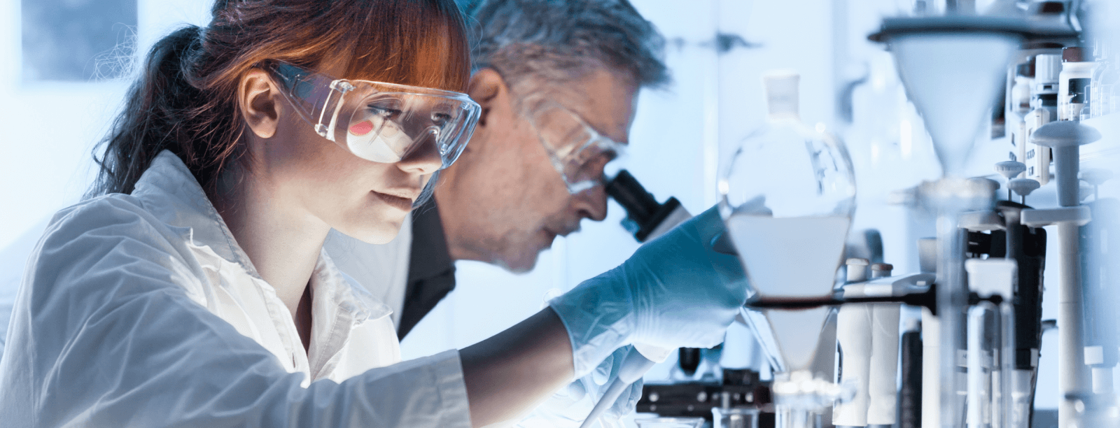 Dos científicos en un laboratorio. La mujer en primer plano lleva gafas de seguridad y guantes y observa un matraz de vidrio
