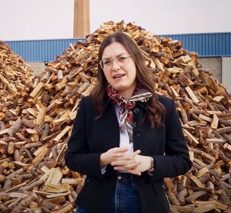 Mujer con gafas y una americana azul, una bufanda roja, una camisa blanca y unos pantalones vaqueros, está delante de una pila de troncos de madera.