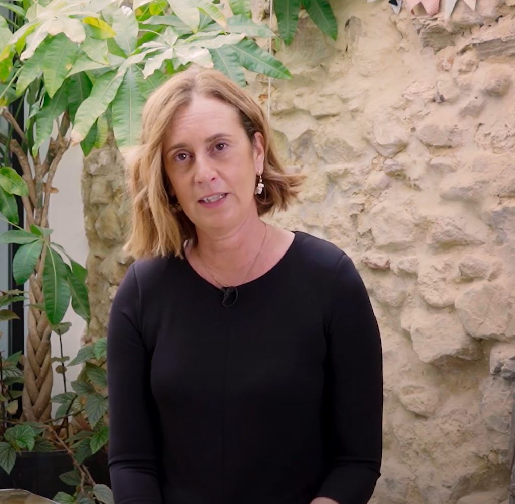 Mujer rubia con jersey negro está conversando delante de una pared de roca con plantas alrededor 