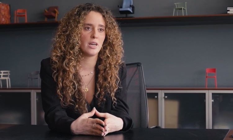 Mujer con pelo rizado y traje negro conversa sentada en una mesa de despacho
