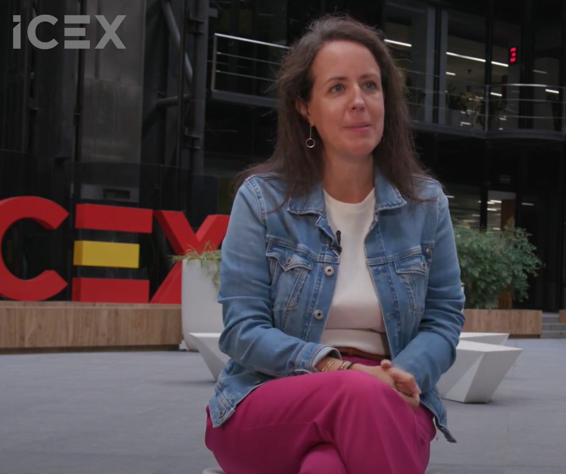 Mujer de pelo castaño con chaqueta vaquera, pantalones rosas y jersey blanco, está sentada en el lucernario de ICEX