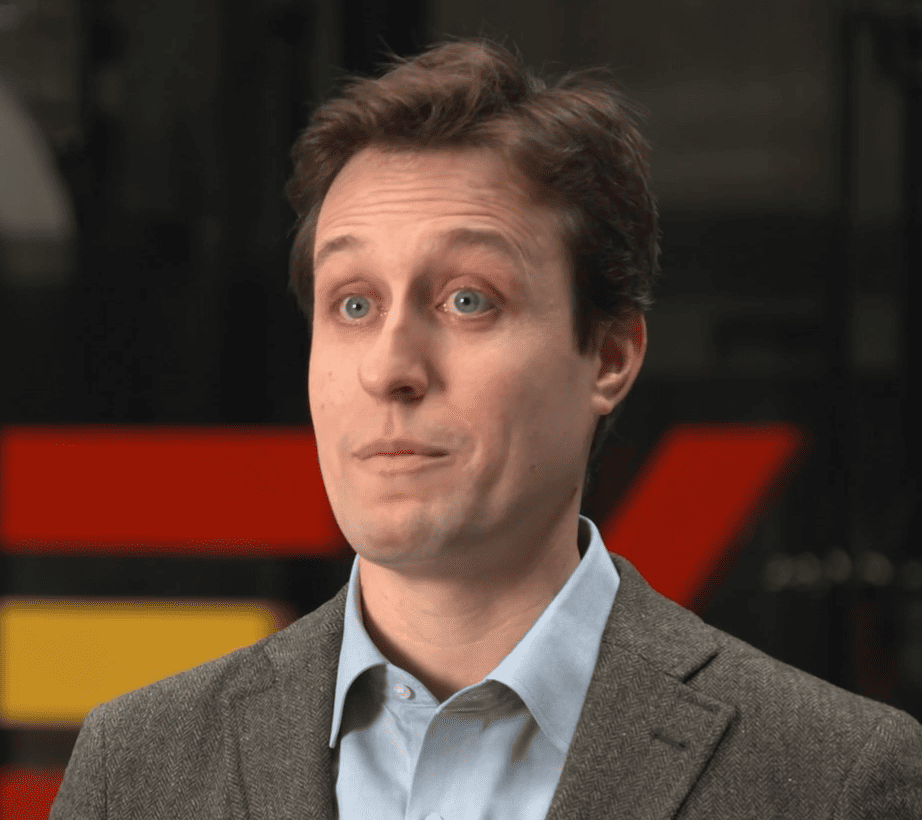  primer plano de una persona con el pelo corto, los ojos azules, una chaqueta marrón y una camisa azul