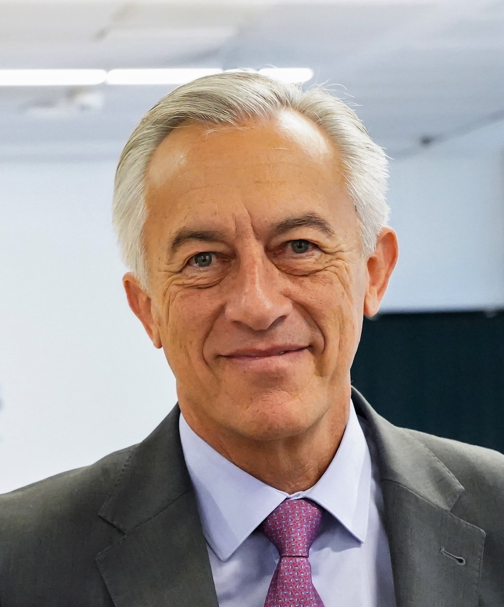 Hombre de cabello gris peinado hacia un lado, viste traje oscuro, camisa clara y corbata rosada. Mira de frente con leve sonrisa.