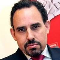 Hombre con cabello oscuro y barba corta tipo perilla. Lleva traje oscuro, camisa blanca y corbata roja. Expresión seria, mirando a cámara.