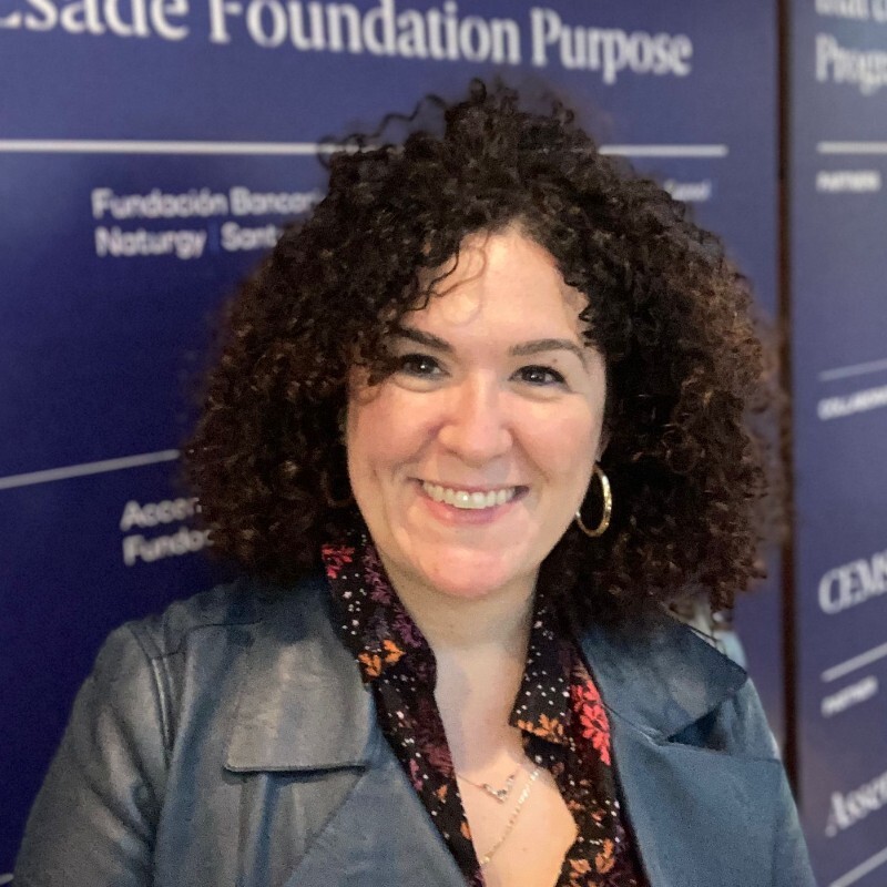 Mujer con cabello rizado oscuro hasta los hombros. Sonríe ampliamente. Lleva chaqueta oscura y blusa estampada.