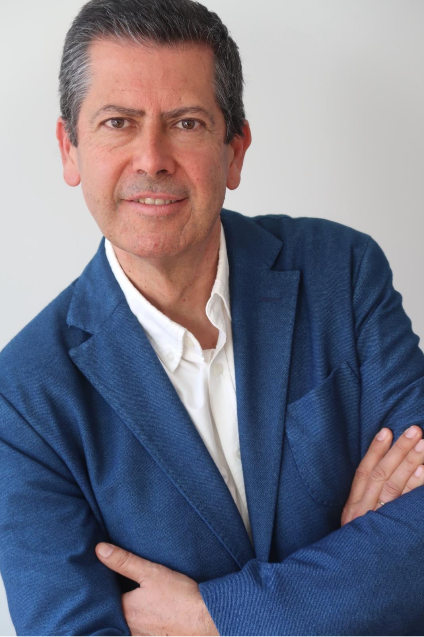 Hombre de cabello corto gris, brazos cruzados. Viste chaqueta azul y camisa blanca sin corbata. Sonríe suavemente