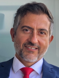 Hombre con cabello corto oscuro y barba corta. Lleva traje azul, camisa blanca y corbata roja. Sonríe levemente.