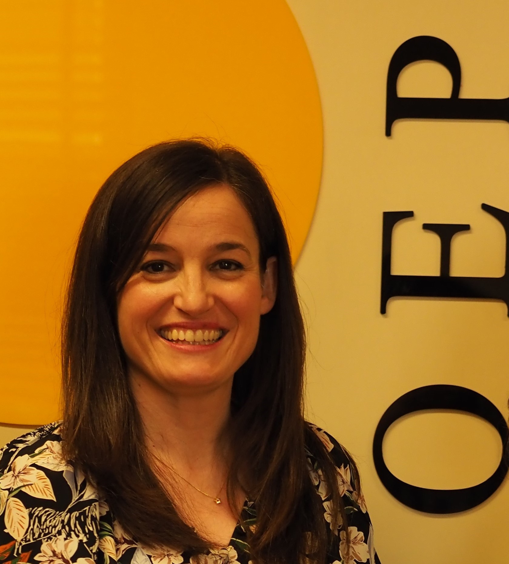 Mujer de cabello liso castaño, a la altura de los hombros. Sonríe mostrando los dientes. Viste blusa con estampado floral.