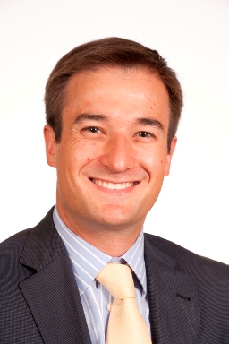 Hombre joven de cabello corto oscuro. Sonríe. Lleva traje oscuro, camisa clara y corbata beige.