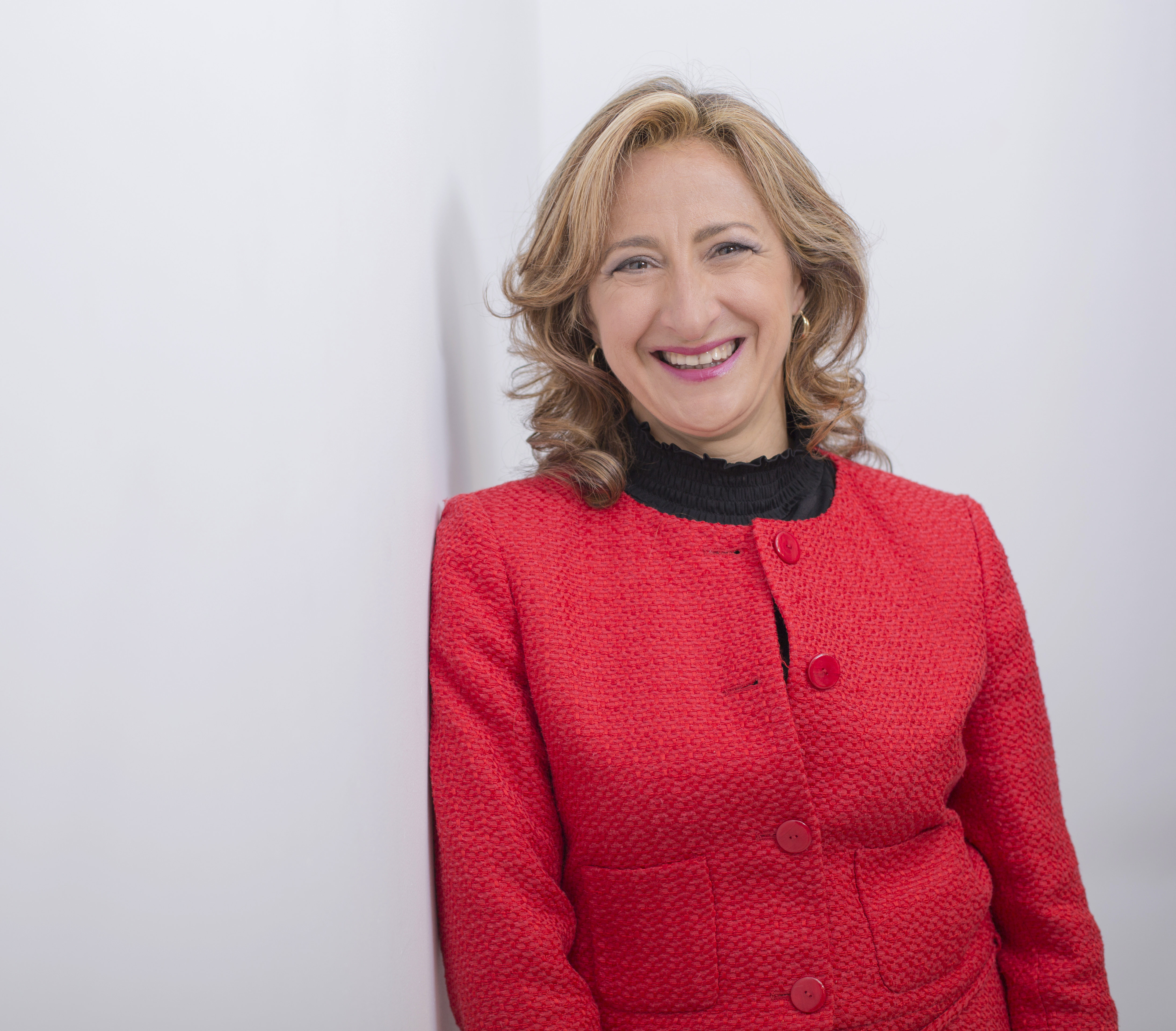 Mujer con cabello claro ondulado hasta los hombros. Sonríe. Lleva chaqueta roja texturizada sobre blusa negra.