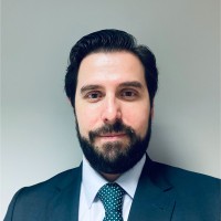 Retrato profesional de una persona con cabello oscuro y barba bien cuidada, vestida con traje oscuro, camisa clara y corbata estampada. El fondo es neutro y la imagen transmite una apariencia corporativa y formal.