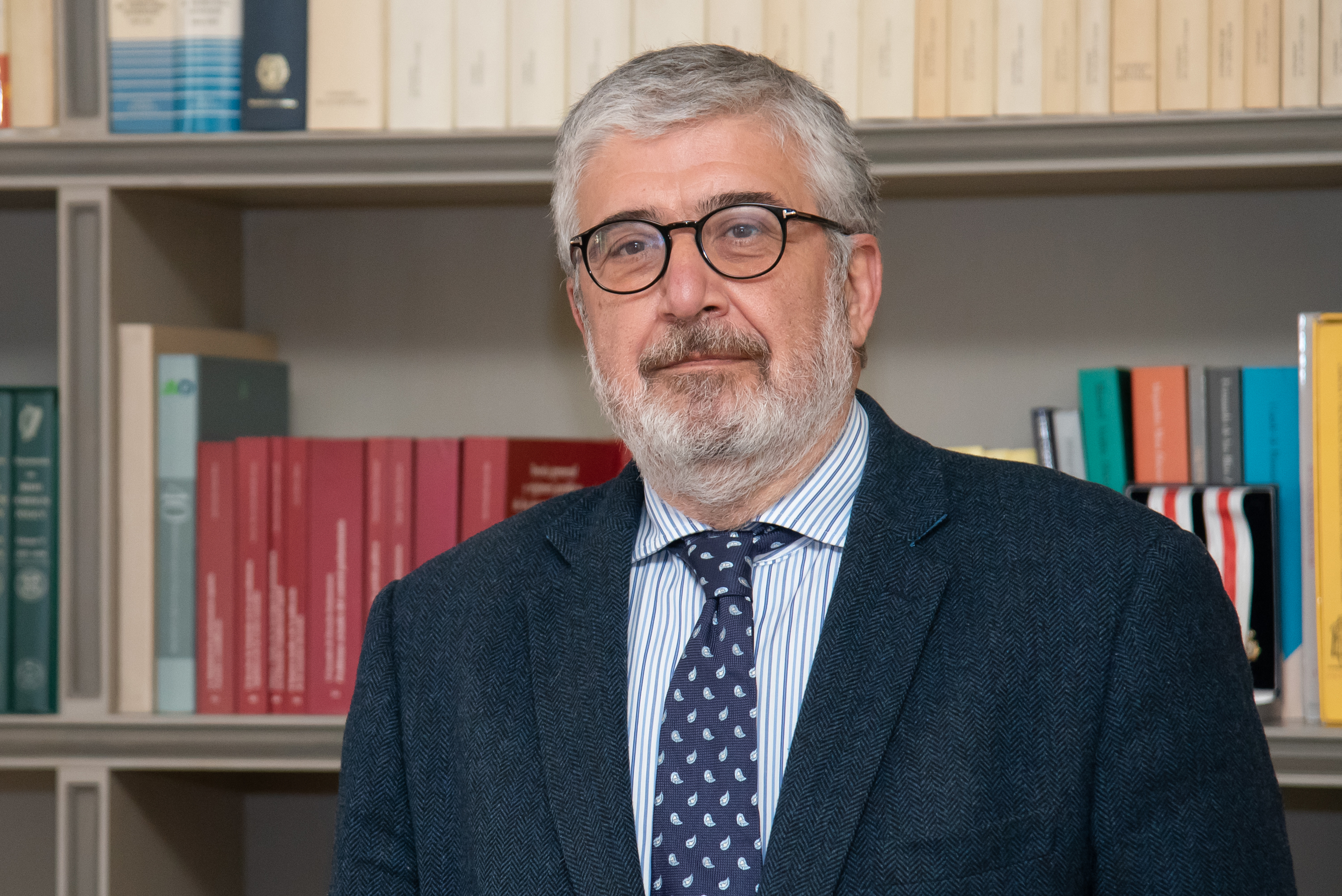 Hombre con cabello corto gris y barba canosa. Lleva gafas redondas, chaqueta oscura, camisa a rayas y corbata azul con pequeños dibujos. Mira de frente con expresión seria. Detrás hay estanterías llenas de libros.