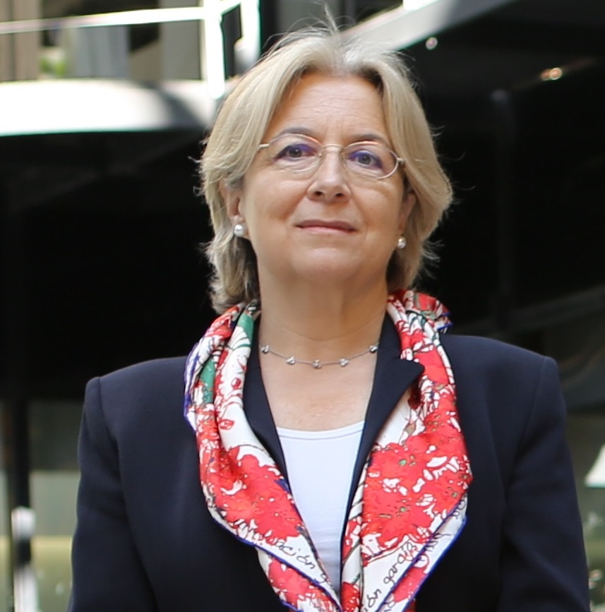 Mujer con cabello corto rubio y gafas finas. Viste chaqueta oscura, blusa clara y un pañuelo rojo con estampado llamativo al cuello. Lleva pendientes pequeños.