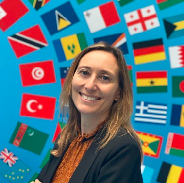 Mujer sonriente con cabello largo castaño claro, suelto sobre los hombros. Lleva chaqueta negra y blusa marrón. Está de pie con los brazos cruzados.