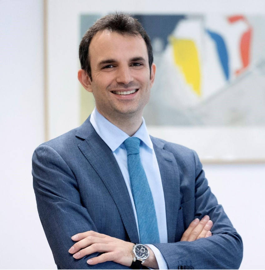 Persona con pelo corto y sonriente con traje  y corbata azul con los brazos cruzados