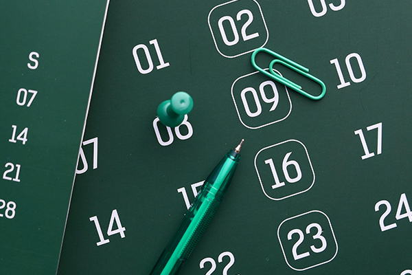 Un calendario de fondo verde oscuro señalando varios días en blanco. Encima hay una chincheta, un bolígrafo y un clip verdes