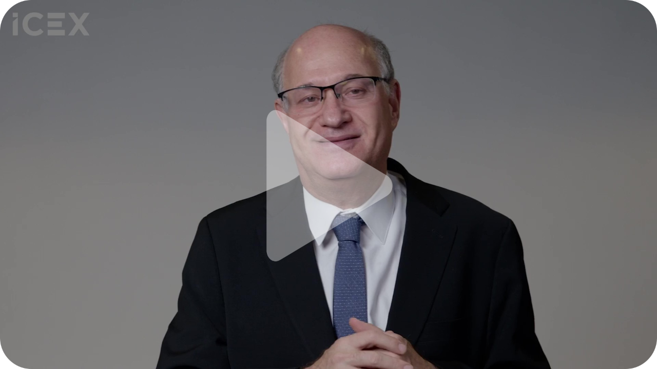 Primer plano de hombre con gafas, chaqueta negra, camisa blanca y corbata azul sobre fondo gris