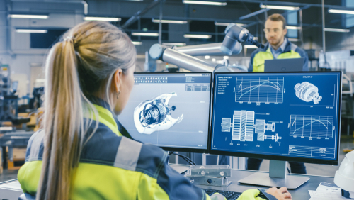 At the Factory: Female Mechanical Engineer Designs 3D Engine on Her Personal Computer. In the Background Male Automation Engineer who Uses Laptop for Programming Robotic Arm.