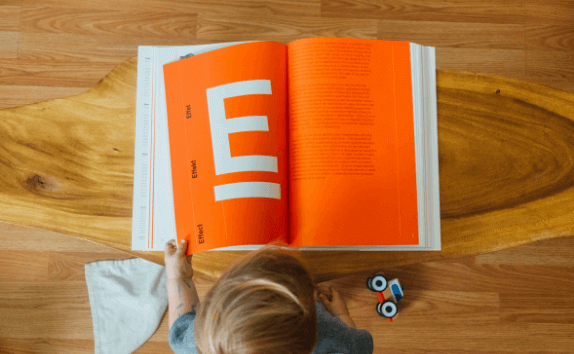Una persona ojea un libro, leyendo dos páginas naranjas. Hay una letra E blanca grande en la página izquierda