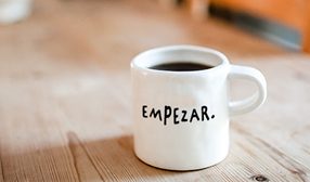 Una taza blanca de café con la palabra empezar escrita en negro. Está situada sobre una superficie de madera