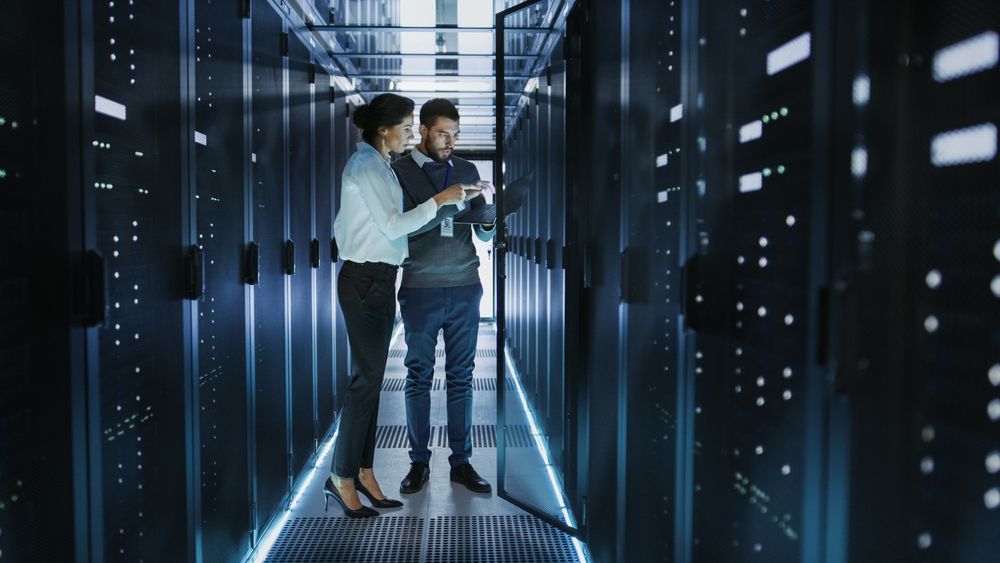 Female IT Technician and Male Server Engineer Talk and Discuss Settings of a Working Data Center. Man Holds Laptop.