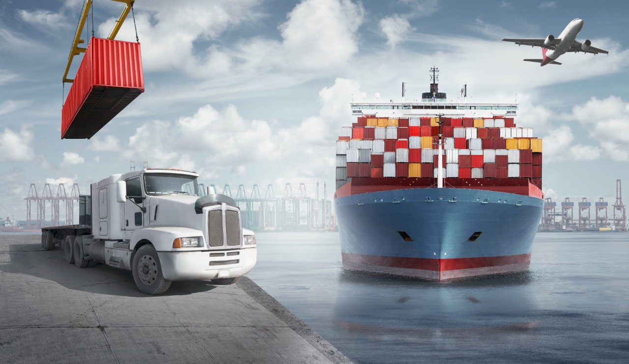 Una grúa coloca un contenedor marítimo en un camión. A la derecha, un buque transporta muchos contenedores