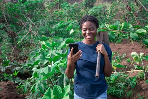 Una mujer africana con una azada en una mano observa el móvil que tiene en la otra mano