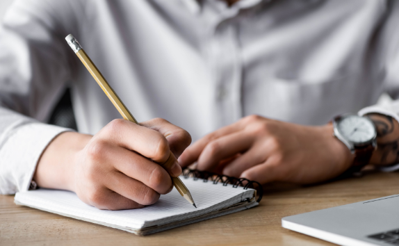 Una persona con camisa gris escribe notas en un cuaderno.