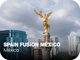 A la izquierda un edificio con fachada acristalada, en el centro una torre con una estatua de un ángel dorado encima, a la derecha varios edificios de oficinas. Spain Fusion México.